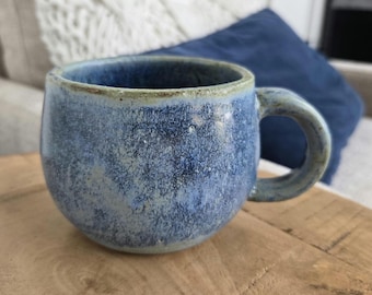 May include: A handmade ceramic mug with a rounded body and a sturdy handle. The mug features a speckled blue glaze with variations in color, creating a textured appearance. The rim has a contrasting light brown color. This mug is perfect for enjoying coffee or tea.