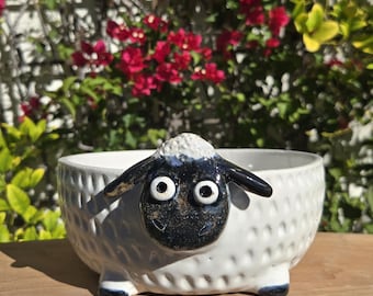 May include: A white ceramic bowl with a sheep face design. The sheep has a black face, ears, and feet, with large, round eyes. The bowl has a textured exterior and is sitting on a wooden surface.