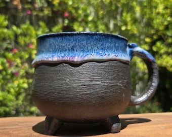 May include: A ceramic mug with a rounded body and three small feet. The lower portion has a matte black textured finish, while the upper rim features a glossy, vibrant blue glaze that drips slightly over the black surface. It has a curved handle.