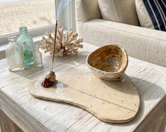 May include: A ceramic incense holder with a beige, organic shape, featuring a small bowl and a place for an incense stick. Smoke rises from the incense. The holder sits on a white wooden table, with a beach scene visible in the background.