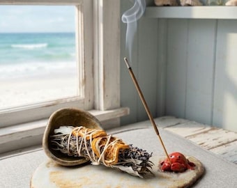May include: A ceramic incense holder featuring a small red crab figure, a bundle of dried sage and lavender tied with twine and orange slices, and a thin incense stick emitting a trail of smoke, all resting on a flat, irregular stone-like base.