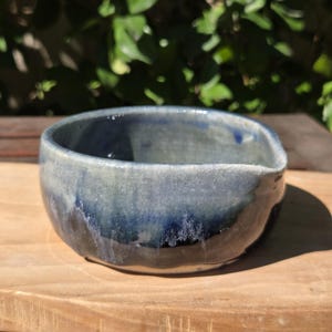 May include: A handmade ceramic bowl with a unique, organic shape. The bowl features a gradient of blue and gray hues, with darker shades at the base and lighter tones towards the rim. The bowl is displayed on a wooden surface.