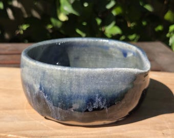 May include: A handmade ceramic bowl with a unique, organic shape. The bowl features a gradient of blue and gray hues, with darker shades at the base and lighter tones towards the rim. The bowl is displayed on a wooden surface.
