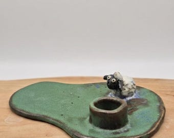 May include: A handmade ceramic candle holder with a green base and a small, black and white sheep figurine. The candle holder has a dark brown rim and a single opening for a candle. The sheep is positioned near the opening.