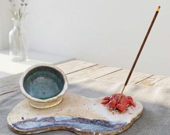 May include: A ceramic incense holder featuring a flat, irregular base with a sandy beige and blue glaze. A small, sculpted red crab sits on the base, holding a lit incense stick. A separate, small, round bowl with a teal interior is attached.