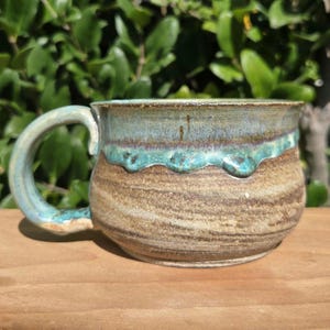 May include: A ceramic mug with a rounded body and a wide handle. The lower portion features a textured, earthy brown and tan marbled finish, while the upper rim is glazed in a vibrant, glossy turquoise blue that drips down the sides.
