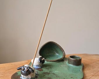 May include: A green ceramic incense holder featuring two sculpted sheep figures with black faces and white bodies. The base has an irregular shape with a small bowl and a cylindrical opening. A single wooden incense stick stands upright in the holder.