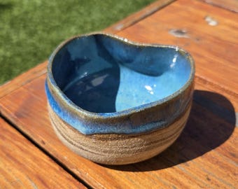 May include: Handmade ceramic bowl with a unique heart-shaped design. The interior is a vibrant blue, contrasting with the earthy, textured exterior. The rim features a gradient of blue and brown hues, adding to its artisanal charm.