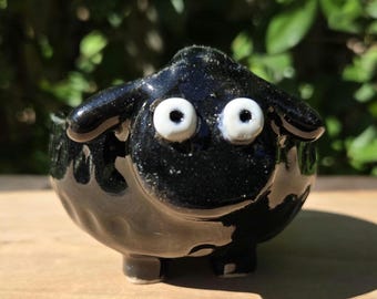 May include: A small, black ceramic sheep figurine with white eyes. The sheep has a glossy finish and is sitting on a wooden surface. The figurine is a decorative item.