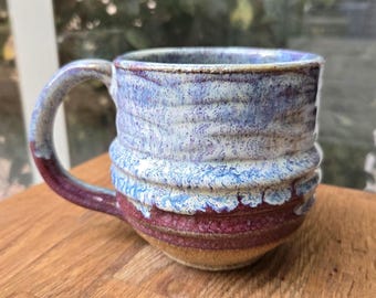 May include: Handmade ceramic mug with a unique glaze. The mug features a marbled effect with shades of blue, white, and purple. The base is a deep burgundy color, and the handle is a matching burgundy. The mug is designed for hot beverages.