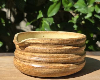 May include: A handmade, ceramic bowl with a light brown glaze. The bowl has a textured, ridged exterior and a smooth, light yellow interior. The bowl has a small spout and sits on a wooden surface.