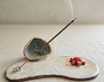 May include: A ceramic incense holder featuring a flat, off-white base with a small, sculpted red crab and a bowl-shaped depression. A thin incense stick is inserted into a small holder on the base, with a thin trail of smoke rising upward.