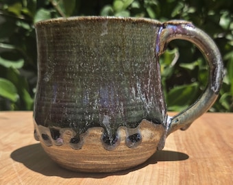 May include: A ceramic mug featuring a dark, mottled green and brown glaze on the upper portion that drips down over a natural, unglazed tan clay base. The mug has a sturdy, curved handle and a cylindrical body with a slightly rounded bottom.