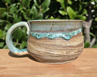 May include: A handmade ceramic mug with a rounded body and a large handle. The mug features a brown base with blue and turquoise accents around the rim and handle. The mug is sitting on a wooden surface.