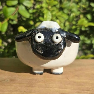 May include: A small ceramic figurine shaped like a sheep with a glossy black face, two prominent white eyes, and a white body. The sheep has wide-set eyes and a smooth, rounded form, standing on four short, stubby legs with black-tipped feet.
