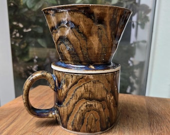 May include: A handmade ceramic coffee pour-over set. The mug and pour-over have a brown wood grain pattern with blue accents. The mug has a handle and the pour-over sits on top. The set is on a wooden surface.