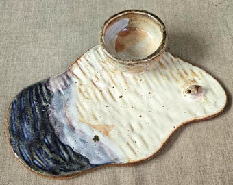 May include: Handmade ceramic serving dish with a small bowl attached. The dish is off-white with textured lines and a dark blue and gray painted section. The small bowl is brown and tan.