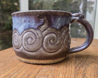 May include: Handmade ceramic mug with a unique swirl design. The mug features a dark brown base, transitioning to a lighter brown with a blue and purple rim. The handle is a matching dark brown. A beautiful piece for coffee or tea.