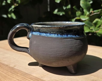 May include: A round, dark gray ceramic mug with a textured, matte finish and three small feet. The upper rim features a glossy, deep blue and black drip glaze. It has a curved handle on the left side and a wide, open top opening.