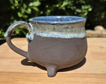 May include: A ceramic mug with a matte dark brown base and three small feet. The upper portion features a glossy, textured glaze in shades of blue, cream, and light green that drips down toward the center. It has a curved handle on the side.