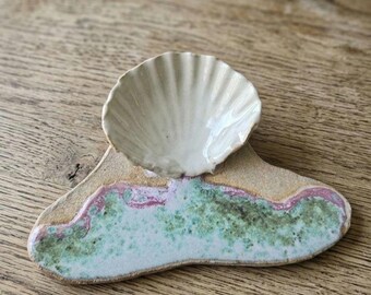 May include: A ceramic trinket dish featuring a sculpted, off-white, ribbed seashell attached to a flat, irregular base. The base has a textured, speckled glaze in shades of light green, white, and a thin border of muted pink along the edge.