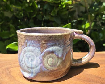 May include: Handmade ceramic mug with a unique spiral design. The mug features a brown base with blue, purple, and green accents. The handle is rounded and attached to the side of the mug. The mug is sitting on a wooden surface.