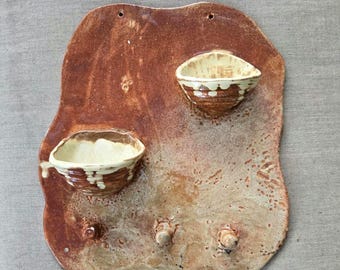 May include: Handmade ceramic wall hanging with two small, bowl-shaped shelves and two pegs. The piece is a mottled brown with cream-colored accents on the shelves. The wall hanging has a rustic, organic aesthetic, perfect for displaying small items.