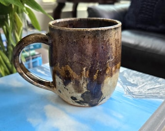 May include: A handmade ceramic mug with a brown and tan glaze. The mug has a rounded shape with a large handle. The glaze has a textured appearance, with darker brown tones at the top and lighter tan tones at the bottom. The mug is sitting on a blue surface.