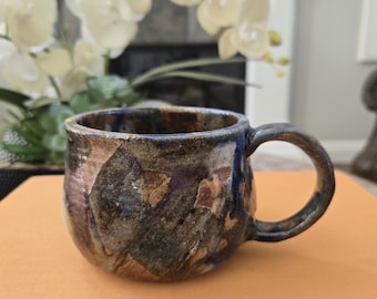 May include: A handmade ceramic mug with a rounded shape and a thick handle. The surface features a mottled, multi-colored glaze with earthy tones of brown, blue, and tan, showing a textured, uneven finish throughout the exterior of the vessel.