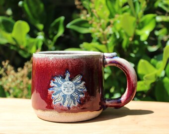 May include: A red ceramic mug with a white rim and handle. The mug has a white and blue sun design with a face on it.