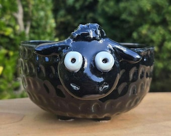 May include: A glossy black ceramic bowl shaped like a sheep, featuring a textured exterior that mimics wool, two protruding ears, and two large, round, white eyes with black pupils centered on the front of the vessel's rounded face.
