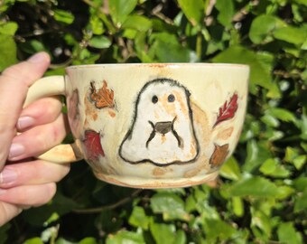May include: Cream-colored ceramic mug with a ghost design and autumn leaf accents. The mug has a handle and is held by a hand against a backdrop of green foliage. The ghost is outlined in black.