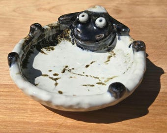 May include: A small, round, handmade ceramic dish featuring a sculpted black sheep head at the rim. The interior is glazed in off-white with dark brown speckled accents, while the exterior rim is textured with small, dark brown rounded protrusions.