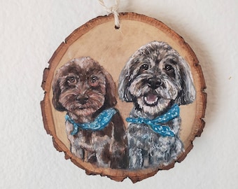 May include: A round wooden ornament featuring a painting of two dogs. The dog on the left is brown, and the dog on the right is gray. Both dogs wear blue bandanas with white polka dots. A natural twine loop is at the top.