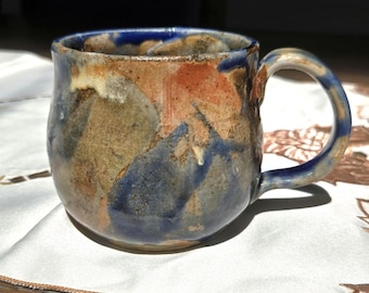 May include: A ceramic mug with a rounded body and a curved handle, featuring a mottled glaze in shades of deep blue, earthy brown, and muted tan. The surface has a textured, uneven appearance with blended color transitions throughout the exterior.