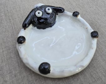 May include: A round ceramic trinket dish featuring a sculpted sheep head at the top with black ears and white eyes. The dish has a glossy white interior and four small black circular accents along the rim, resembling the sheep's hooves.