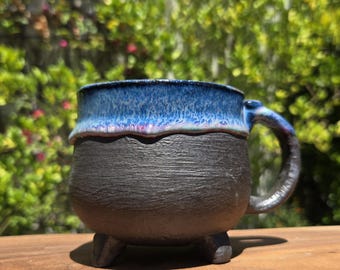 May include: A handmade ceramic mug with a unique design. The mug features a black textured base and a blue glazed rim. It has a rounded body, a handle, and three small feet. The mug is sitting on a wooden surface.
