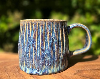 May include: A handmade ceramic mug with a textured, vertical-lined design. The mug features a blend of blue, brown, and gold colors, with a rounded handle. The mug is sitting on a wooden surface.