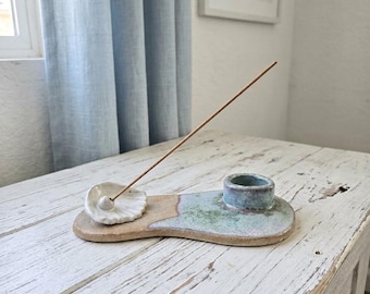 May include: A ceramic incense holder with an irregular, elongated base featuring a tan, unglazed underside and a mottled blue and green glazed top. It includes a small white shell-shaped incense stick holder and a cylindrical candle holder.