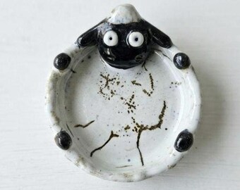 May include: A small, round, white ceramic dish featuring a black sheep head at the top rim. The interior of the dish is speckled with black dots and features a simple black line drawing of a sheep body. Four small black feet are attached to the base.