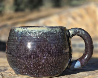 May include: A handmade ceramic mug with a dark purple glaze. The mug has a rounded shape with a large handle and a light blue accent. The rim of the mug has a light green and white color.