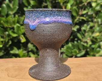 May include: A handmade ceramic goblet with a dark brown body and a flared base. The rim is glazed with a blend of blue and purple hues, creating a striking contrast. The goblet is displayed on a wooden surface, showcasing its unique design and craftsmanship.