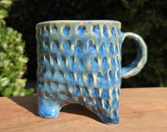 May include: A handmade ceramic mug with a unique textured surface and three small feet. The mug features a blue and cream glaze, with a rounded handle. The mug is sitting on a wooden surface.