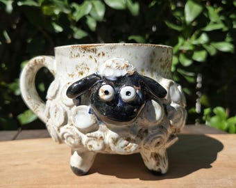 May include: A ceramic mug with a sheep design. The mug is white with a textured surface and a black and white sheep face on the front. The mug has a handle and three small feet. The mug is sitting on a wooden surface.
