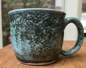 May include: A handmade ceramic mug with a speckled teal and black glaze. The mug has a rounded body, a sturdy handle, and a white rim. The mug is sitting on a wooden surface.