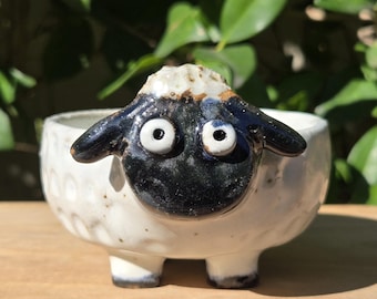 May include: A white ceramic sheep-shaped bowl with a black face and ears. The bowl has a textured exterior and is resting on a wooden surface. The sheep has large, round eyes and small legs.