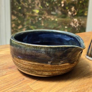 May include: A handmade ceramic bowl with a spout, featuring a blend of blue, brown, and tan hues. The bowl's interior is a deep blue, with a textured, earthy exterior. The design suggests a rustic, artisanal aesthetic, suitable for serving or display.