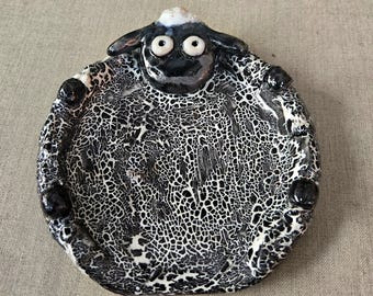 May include: A ceramic sheep-shaped dish with a black and white crackle glaze. The sheep has a black face, ears, and feet, with white eyes and a small white patch on its head. The dish is round with a slightly raised rim.
