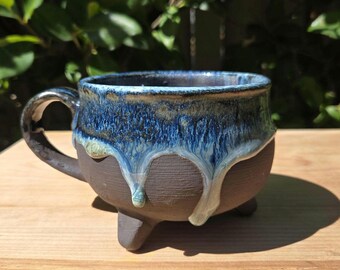 May include: A ceramic mug with a dark brown matte base and a glossy blue glaze dripping from the rim. The mug features a rounded body, a curved handle on the left side, and three short, sturdy feet supporting the base of the vessel.