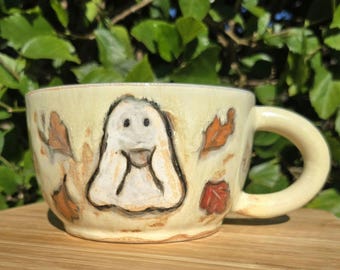 May include: A ceramic mug with a light yellow glaze, featuring a hand-painted ghost design and autumn leaves. The mug has a rounded handle and is set on a wooden surface. The ghost is white with black details.